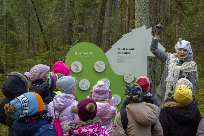 Sieviete kazas maskā pie interaktīvā steda vada nodarbību mazu bērnu pulkam, kas ģēbušies krāsainos apģēbos.